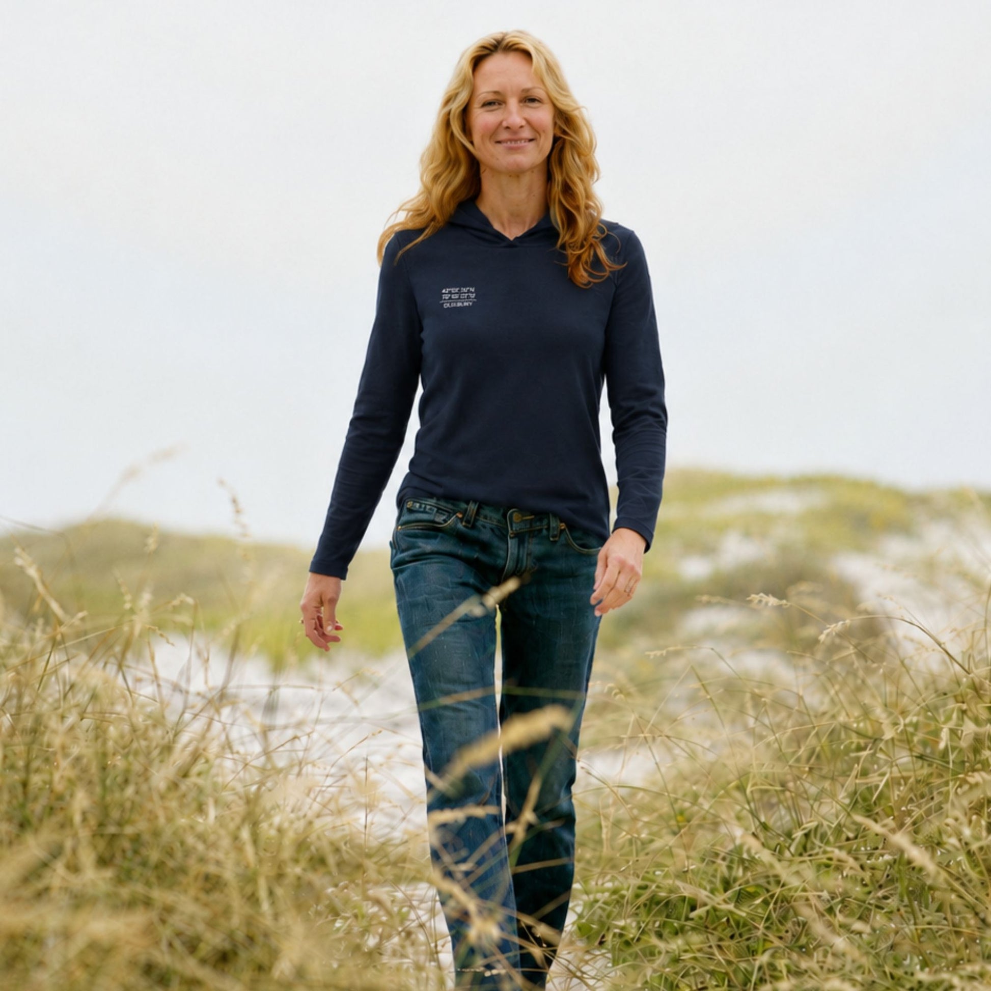 A woman walking outdoors in a grassy field wearing a navy blue long-sleeve quarter-zip pullover with a small white logo on the left chest, paired with blue jeans.