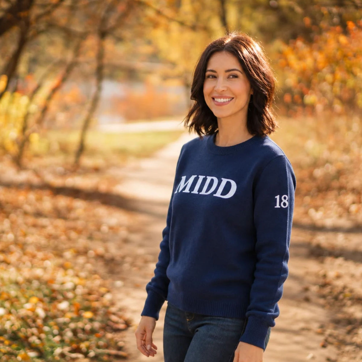 A woman stands on an autumn tree-lined path wearing a navy blue crewneck sweatshirt with 'MIDD' and '18' printed in white on the front and sleeve.
