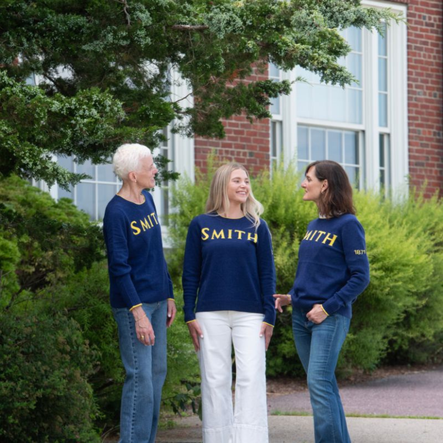 Three women wearing navy 'SMITH' sweatshirts standing outdoors with greenery and a building in the background.