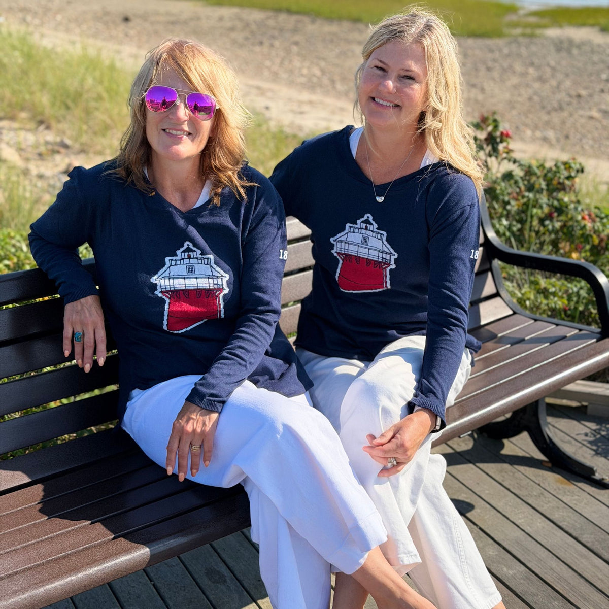 Two women sitting on a wooden beachside bench wearing matching navy blue long-sleeve shirts with a lighthouse graphic on the front, paired with white pants.