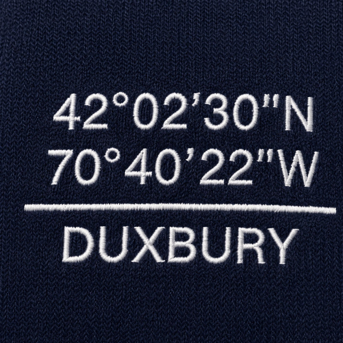 Coordinates of Duxbury, Ma. embroidered in white letters on a navy blue knit sweater
