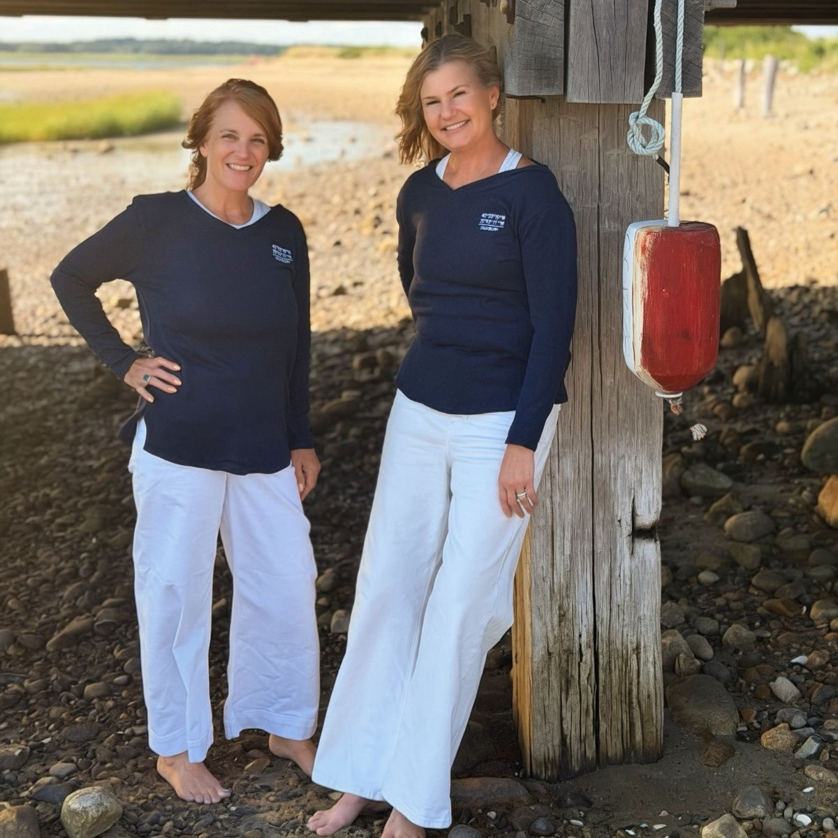 Two women standing outdoors on a rocky shoreline next to a wooden post, wearing matching navy long-sleeve V-neck sweaters with a small white logo on the chest and white wide-leg pants.