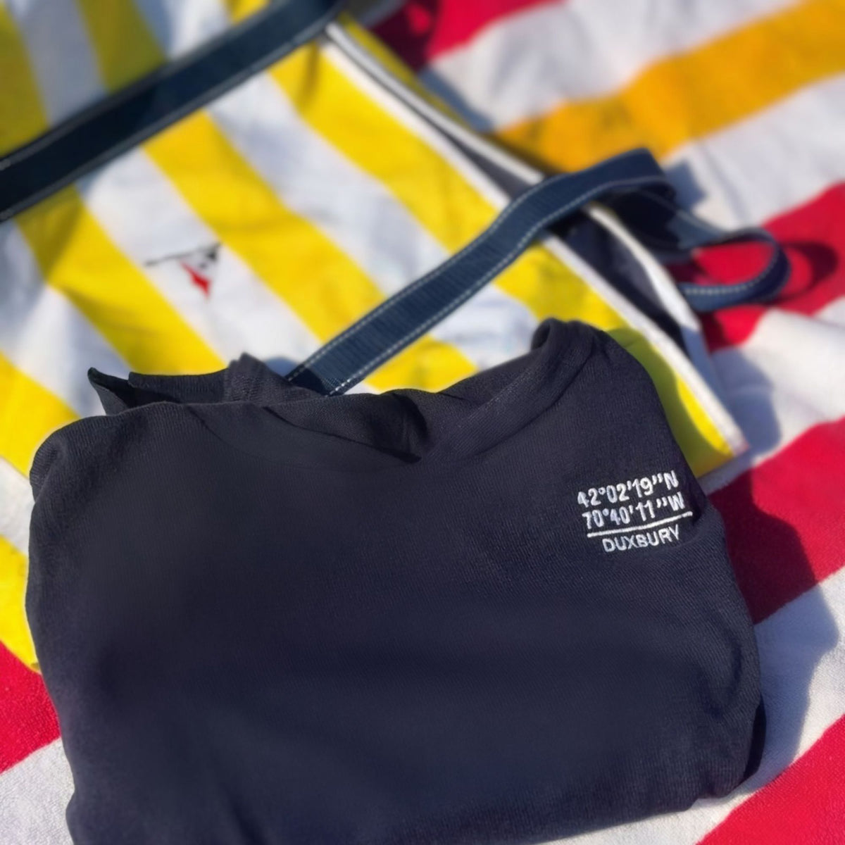 A folded navy blue sweater with white embroidered coordinates and the word 'DUXBURY' lies on a yellow, white, and red striped blanket. A navy blue tote bag is partially visible in the background.