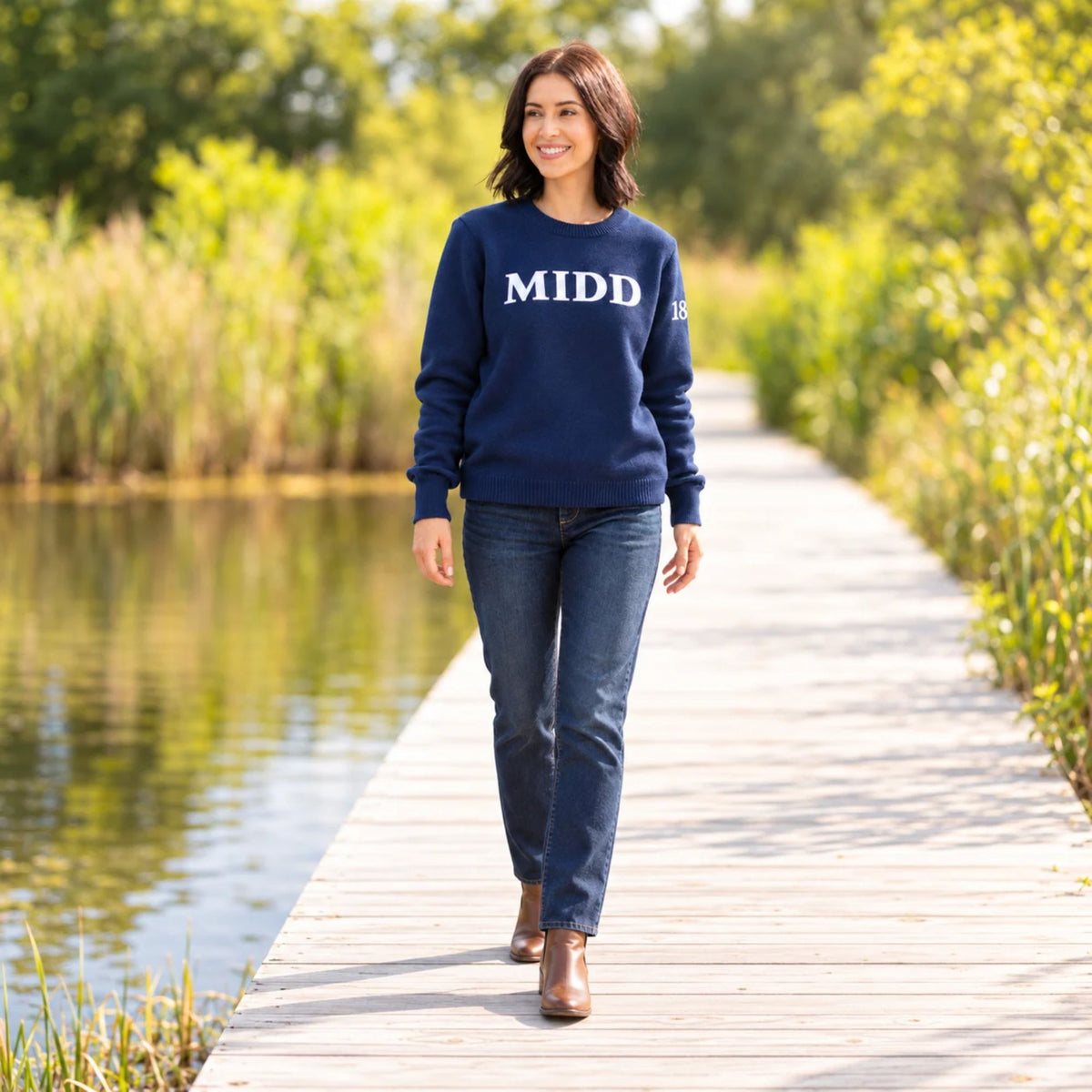 A woman walking on a wooden boardwalk beside a calm lake, wearing a navy blue crewneck sweatshirt with 'MIDD' in white block letters across the chest, paired with dark blue jeans and brown ankle boots.