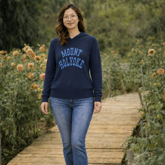 A woman wearing a navy blue hoodie with 'MOUNT HOLYOKE' printed in light blue varsity letters, walking on a wooden boardwalk path surrounded by green foliage and orange flowers.
