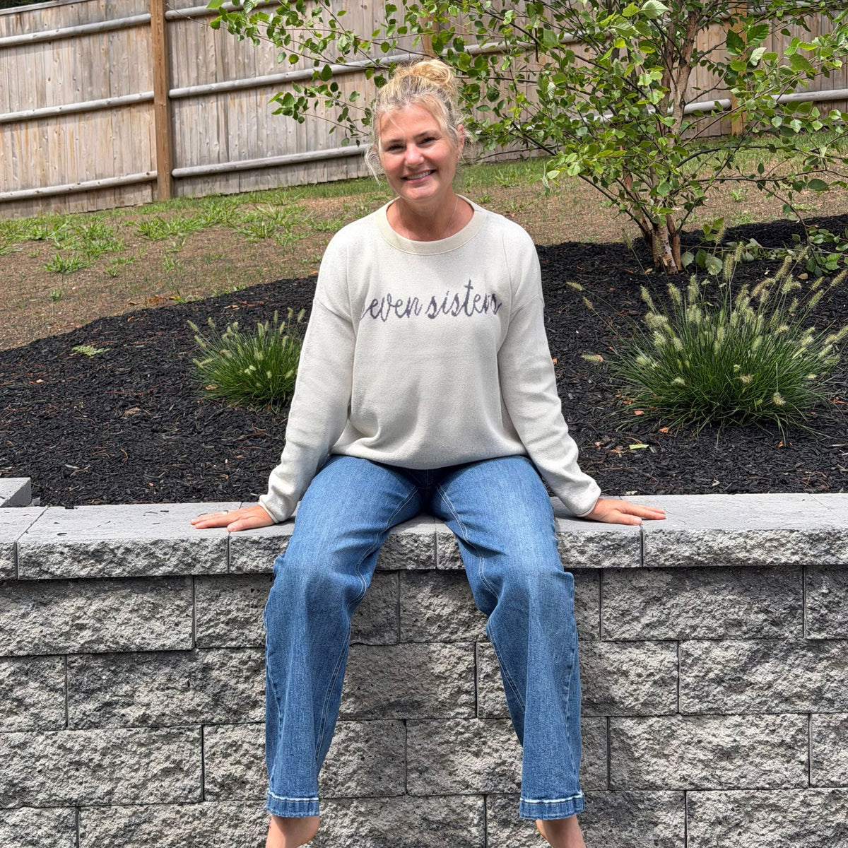 A woman sitting on a gray stone retaining wall in a backyard, wearing a cream sweatshirt with 'seven sisters' written in dark purple script across the chest and blue jeans.