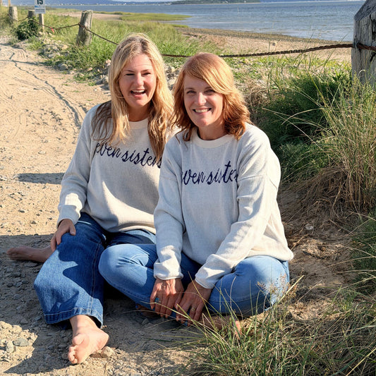 Two women sitting on a sandy beach path wearing matching light gray sweaters with 'seven sisters' written across the front in dark purple script, paired with blue jeans.