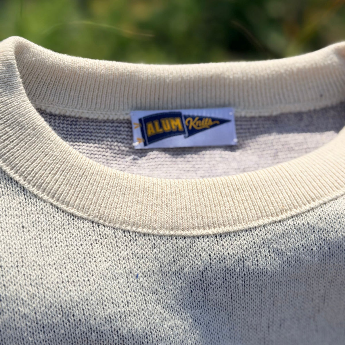 Close-up of a cream-colored knit crewneck sweater with a visible 'ALUMKNITS' neck label, set against a blurred green outdoor background.