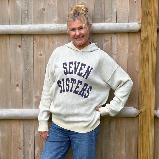 A woman wearing an oversized cream-colored hoodie with 'SEVEN SISTERS' printed in dark navy blue across the chest, standing in front of a wooden fence.