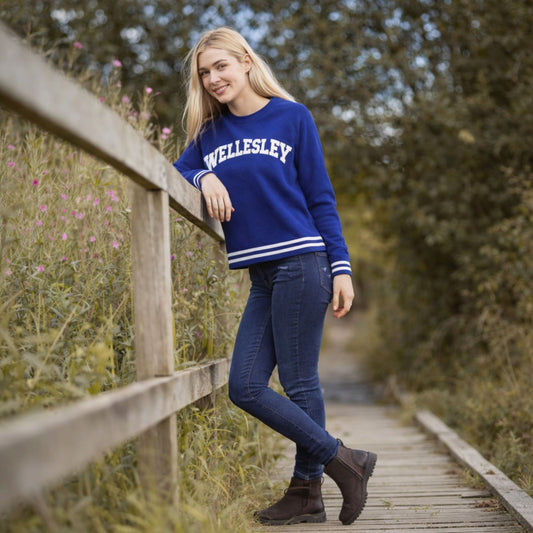 Woman wearing a blue 'Wellesley' sweatshirt on a wooden path in a natural setting