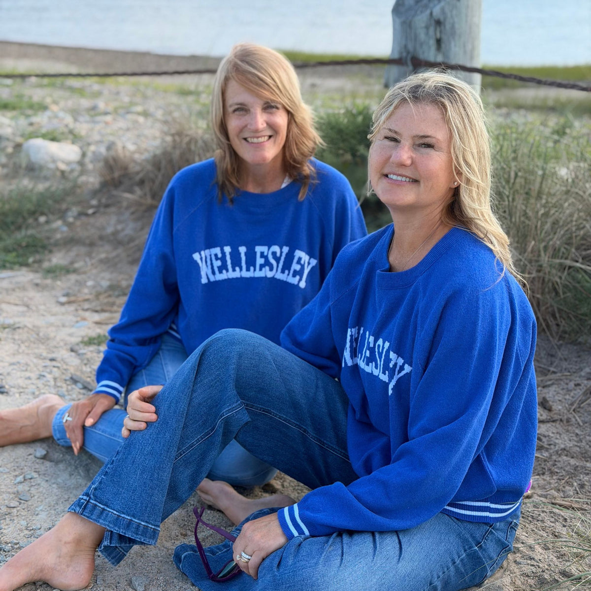 Two women sitting outdoors on sand near water, wearing matching royal blue sweatersd with white 'Wellesley' lettering across the chest.