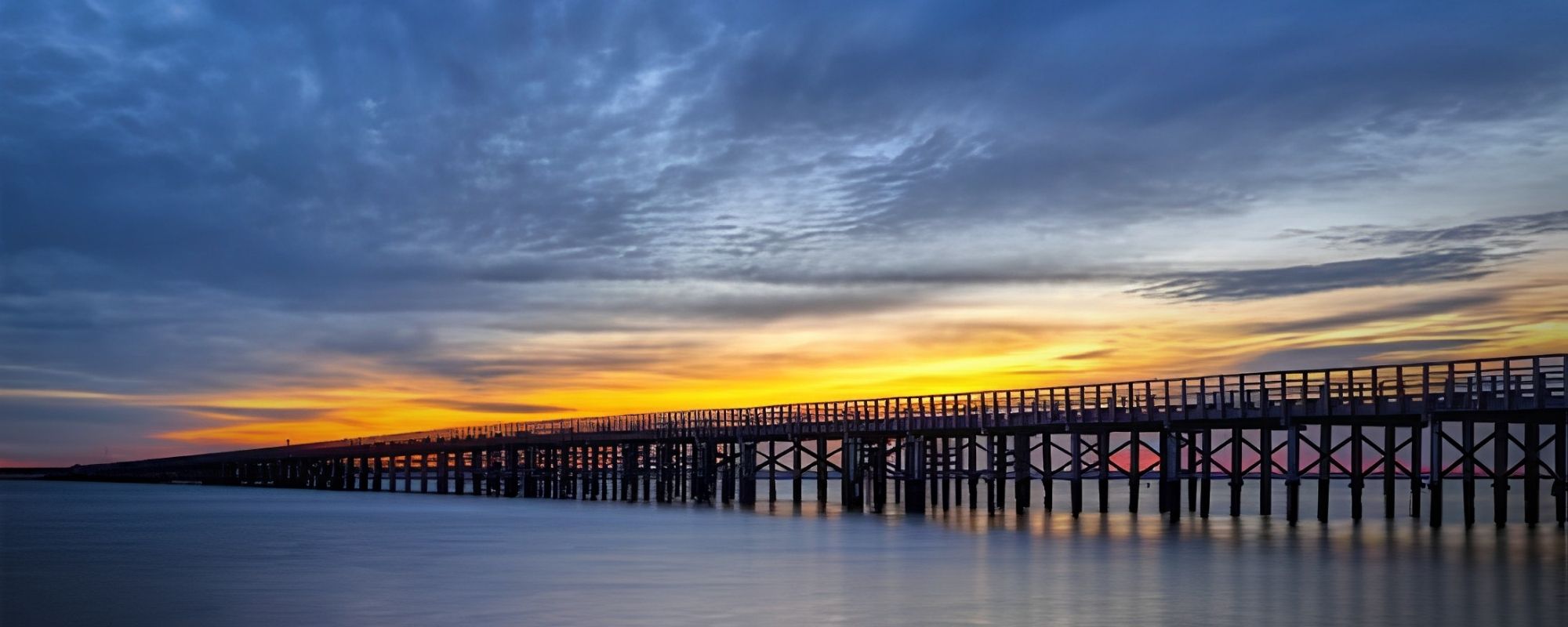 A long wooden pier stretches across calm water under a dramatic sunset sky with blue, gray, and orange clouds.