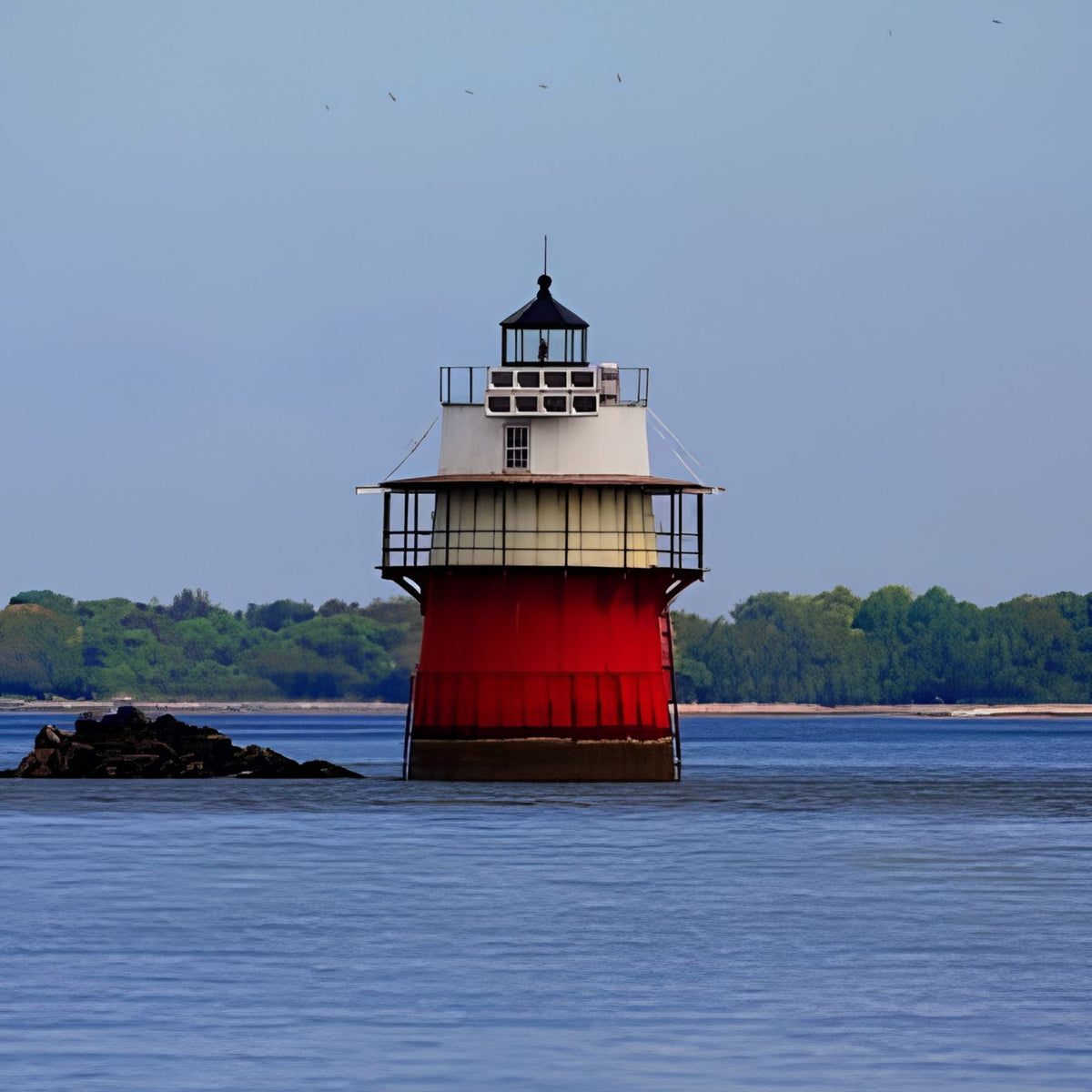 A red and white lighthouse stands in calm blue water under a pale sky, with a tree-lined shore in the background.