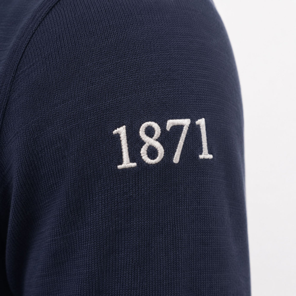 Close-up of a navy blue knit garment sleeve with white embroidered '1871' text on the upper arm, against a light gray background.