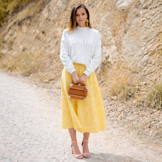 Woman in a white sweater and yellow checkered skirt standing on a dirt road.